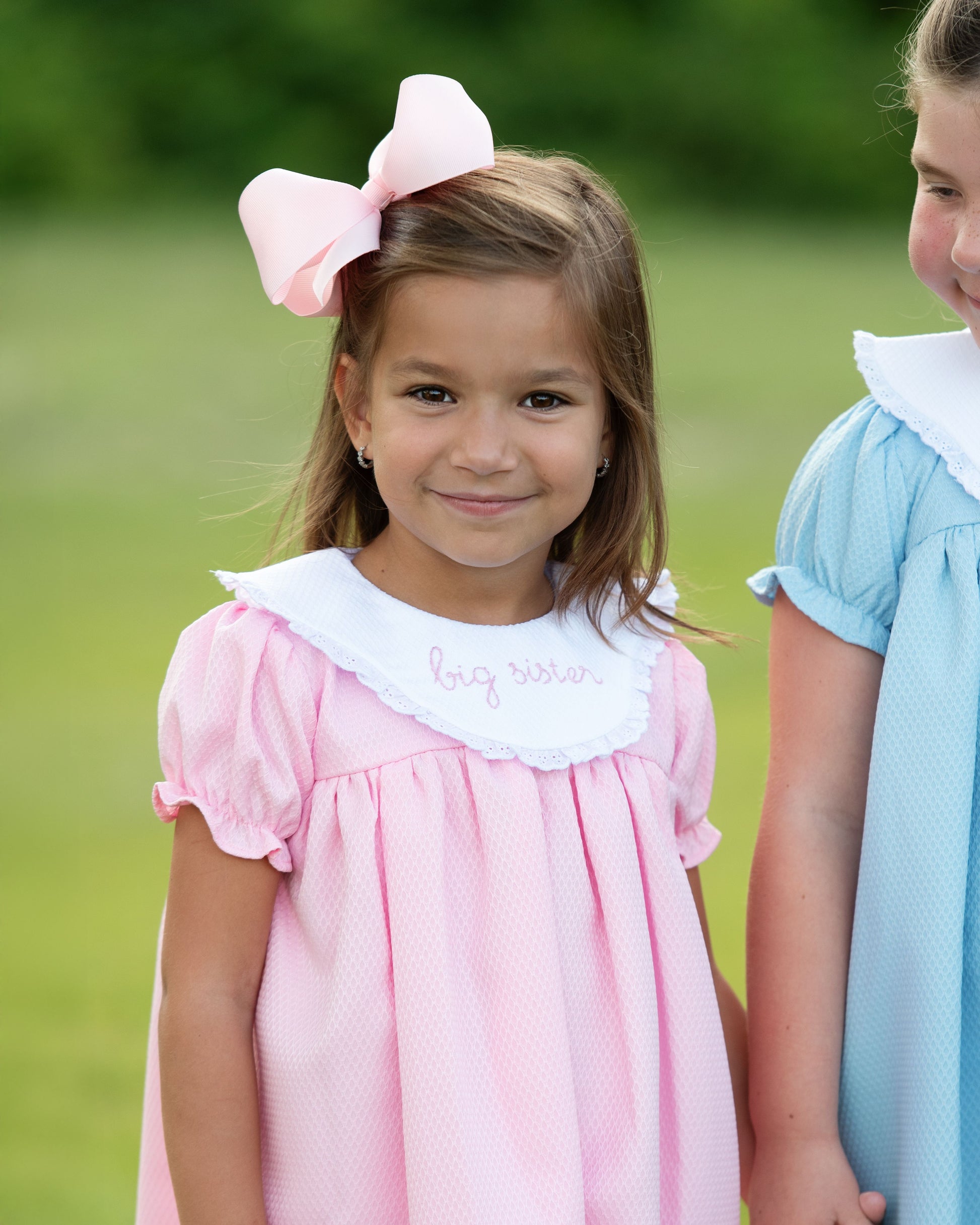 Two young girls standing outdoors, one in a pink dress with a large bow and the other in a blue dress.
