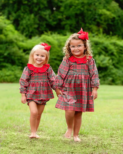 Two young girls walking barefoot on a grassy lawn, wearing matching red and green plaid dresses with red ruffled collars and red bow headbands, with a lush green tree background.