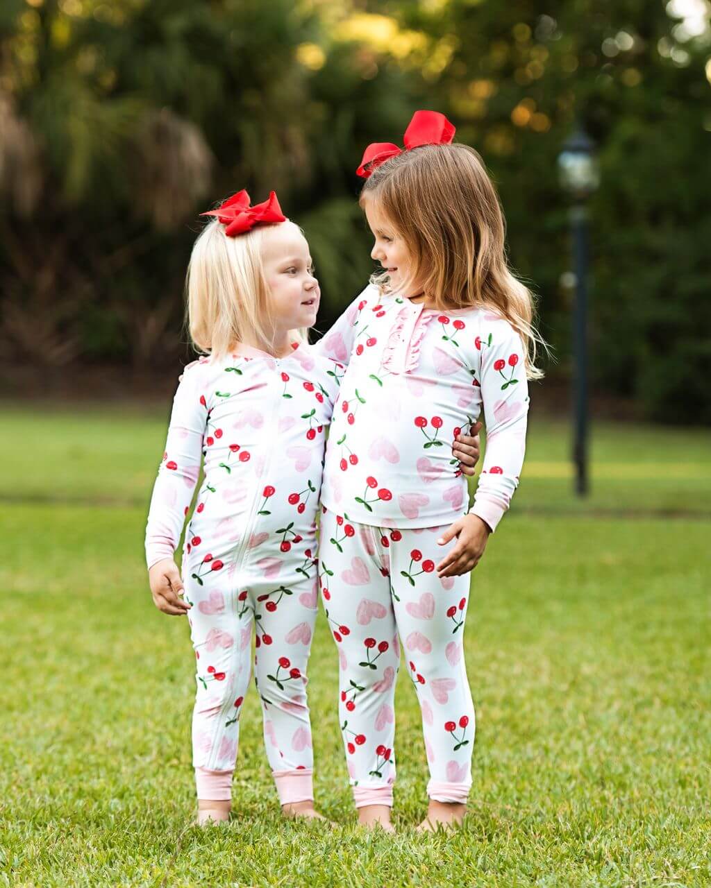 Two young girls wearing cherry-patterned pajamas with a red bow outdoors.