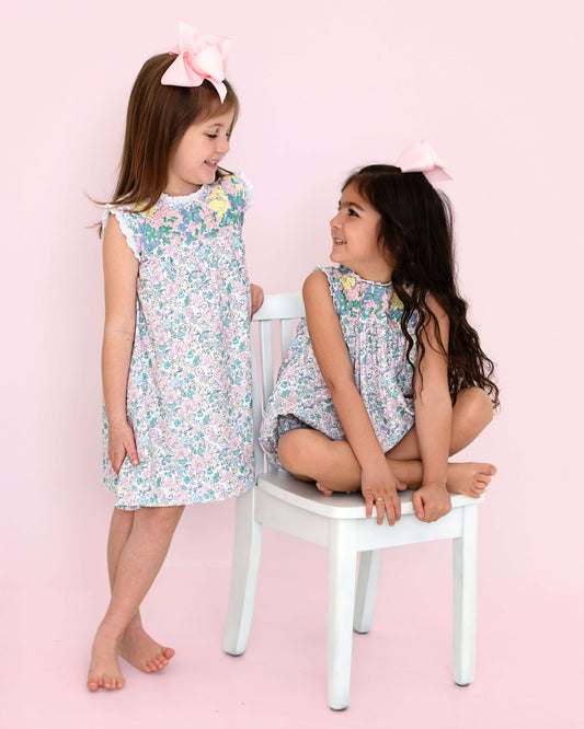 Two young girls wearing floral dresses sitting on a white chair against a pink background
