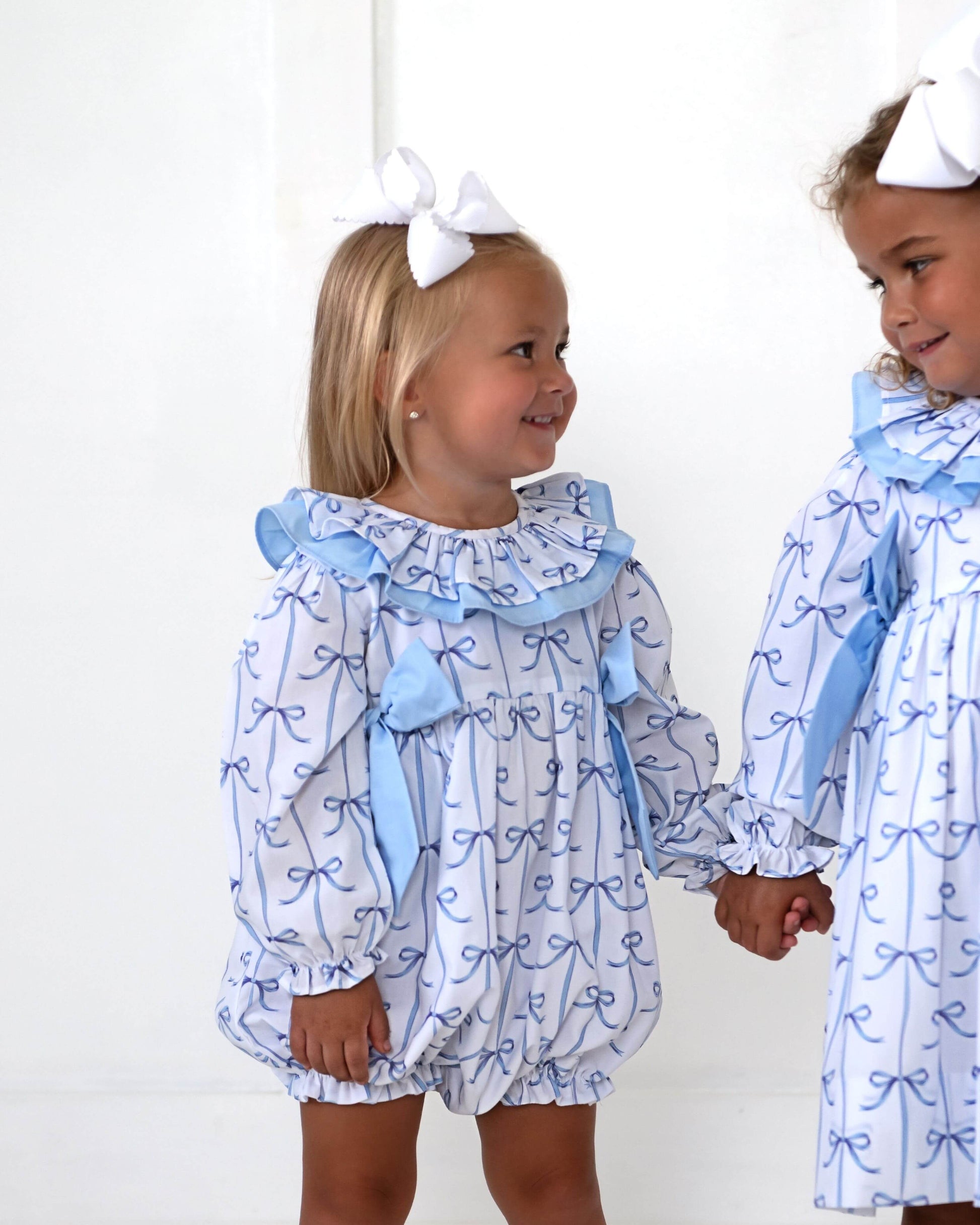 Two young girls wearing matching blue and white outfits with bows, standing against a white background.
