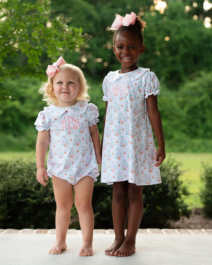 Two young girls wearing matching floral dresses outdoors with greenery in the background.

