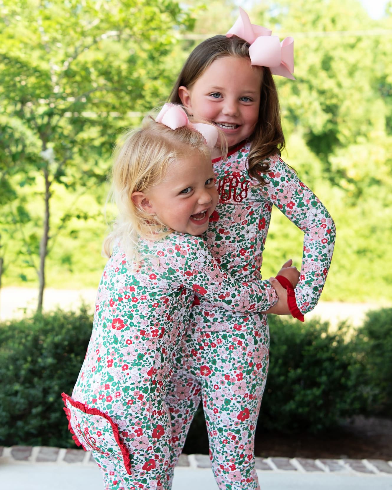 Two young girls wearing matching floral long-sleeve pajamas with red ruffle trim, standing outdoors in front of green trees and hedges. One pajama has a pink monogram on the chest, and both wear pink hair bows.