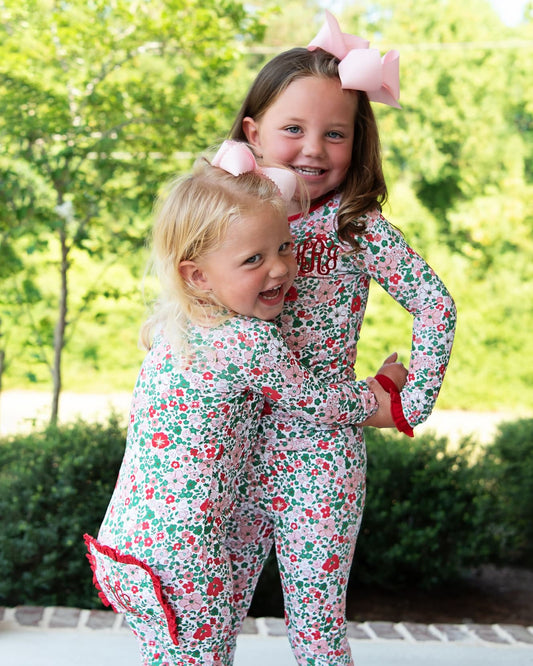 Two young girls wearing matching floral long-sleeve pajamas with red ruffle trim, standing outdoors in front of green trees and hedges. One pajama has a pink monogram on the chest, and both wear pink hair bows.