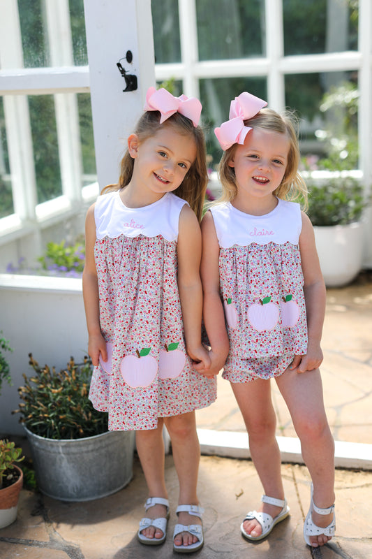 Two young girls wearing matching floral outfits with apples on it and white sandals.