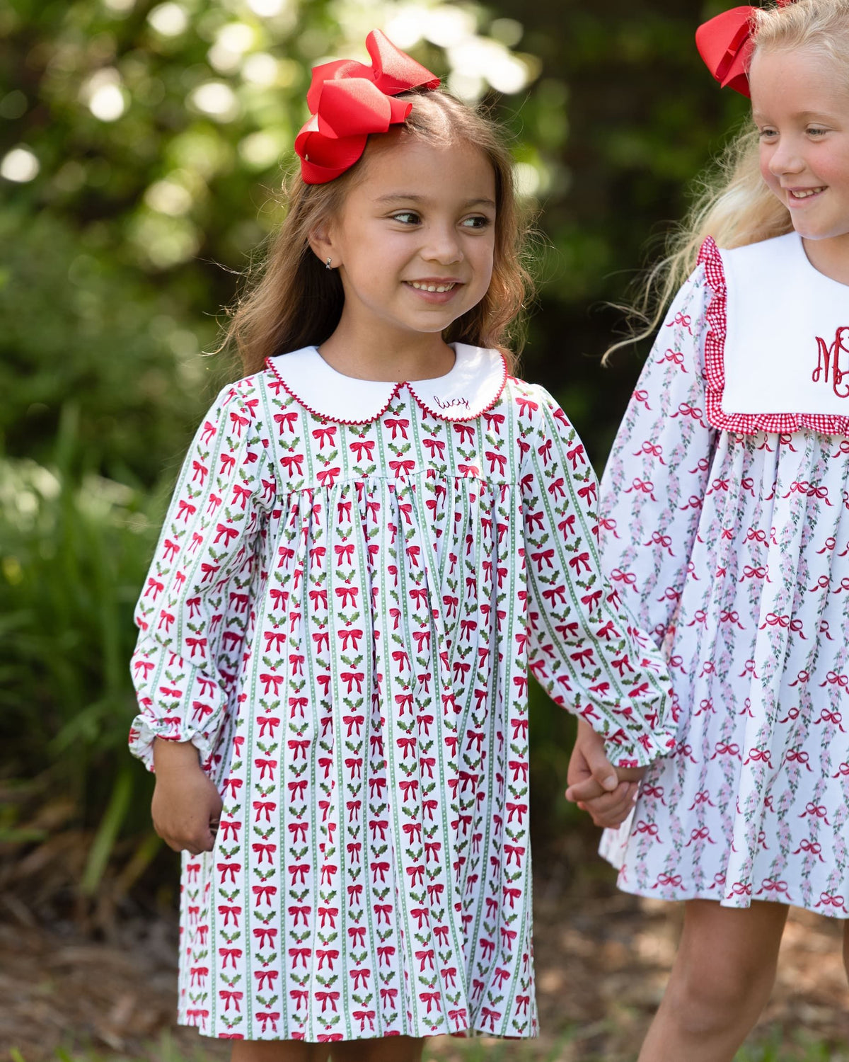 Two young girls wearing matching long-sleeve dresses with a white base and red bow pattern, standing outdoors in a garden setting. The girl on the left wears a large red bow in her hair, and both girls are smiling and holding hands.