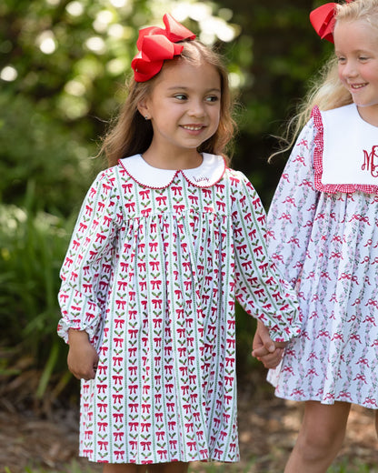Two young girls wearing matching long-sleeve dresses with a white base and red bow pattern, standing outdoors in a garden setting. The girl on the left wears a large red bow in her hair, and both girls are smiling and holding hands.