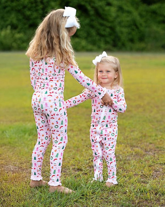 Two young girls wearing matching long-sleeve footed pajamas with a colorful Christmas-themed print, standing on a grassy lawn outdoors.