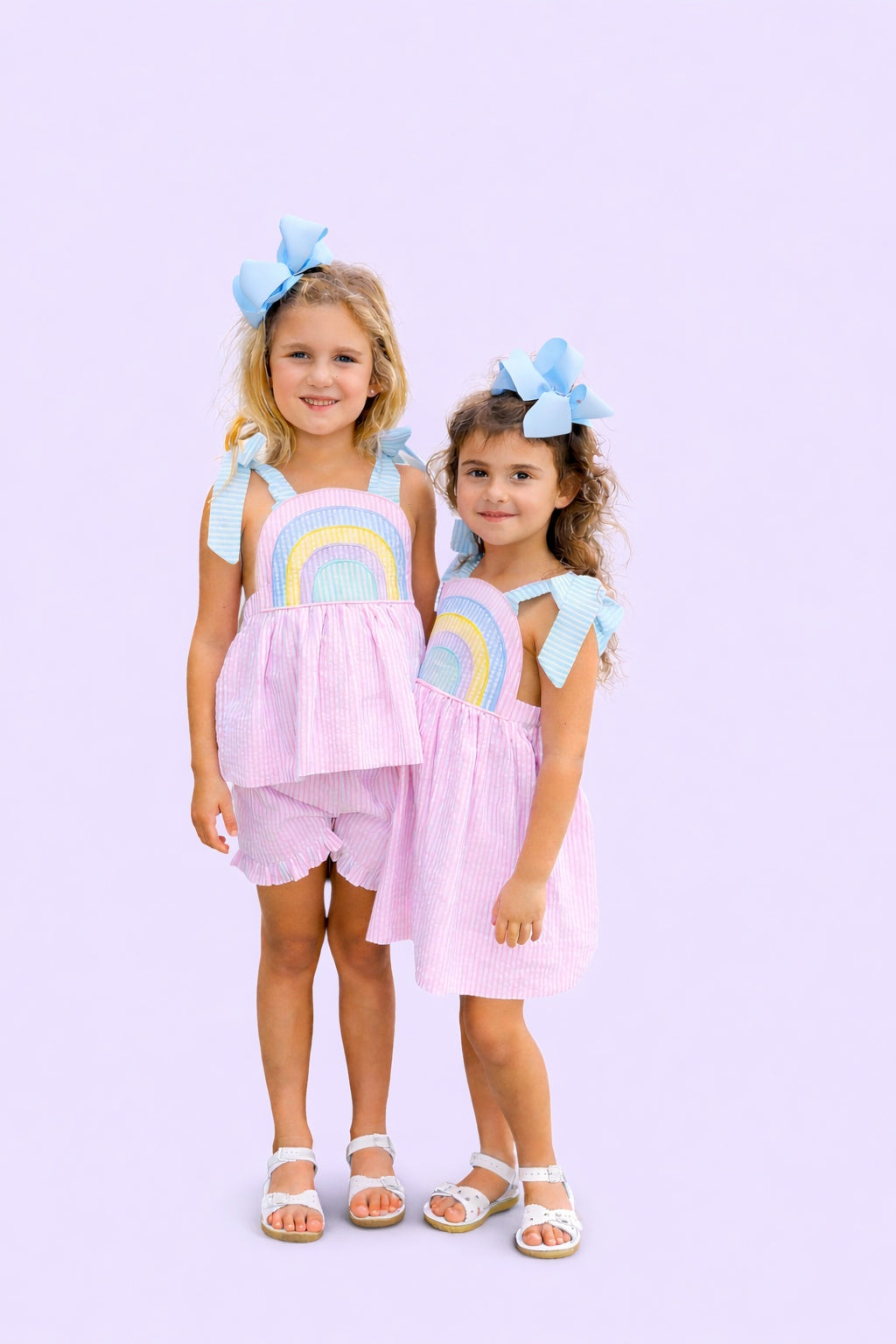 Two young girls wearing matching pink outfits with rainbow designs on a light purple background