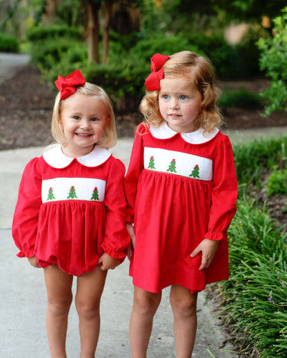 Two young girls wearing matching red dresses with Christmas tree patterns outdoors.