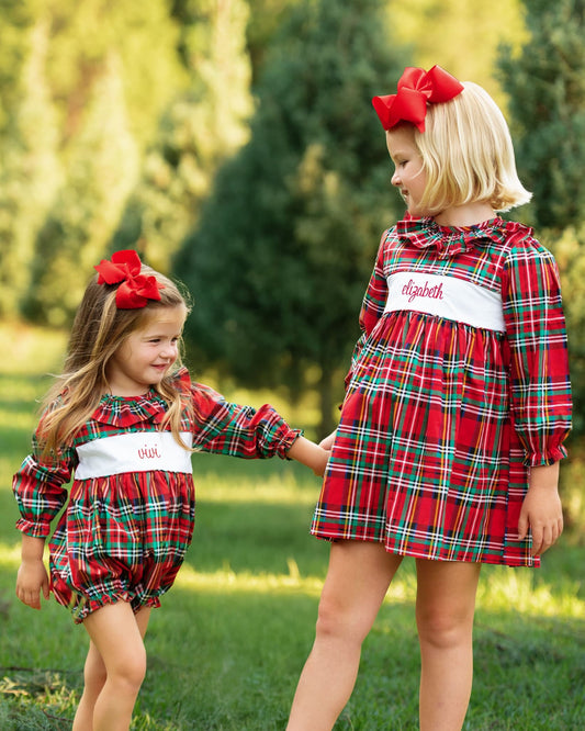 Two young girls wearing matching red plaid dresses and bubble shorts with white waist bands, standing outdoors on grass with trees in the background. One dress has the name 'Elizabeth' embroidered on the front.