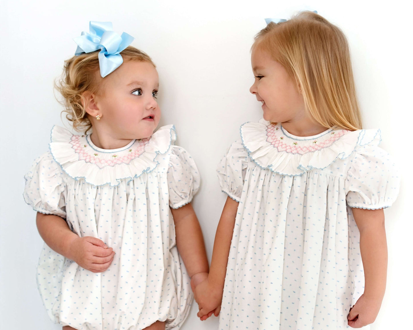 Two young girls wearing matching white dresses with ruffled collars against a plain background.