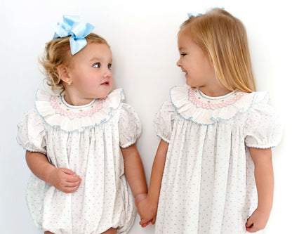 Two young girls wearing matching white dresses with ruffled collars against a plain background.