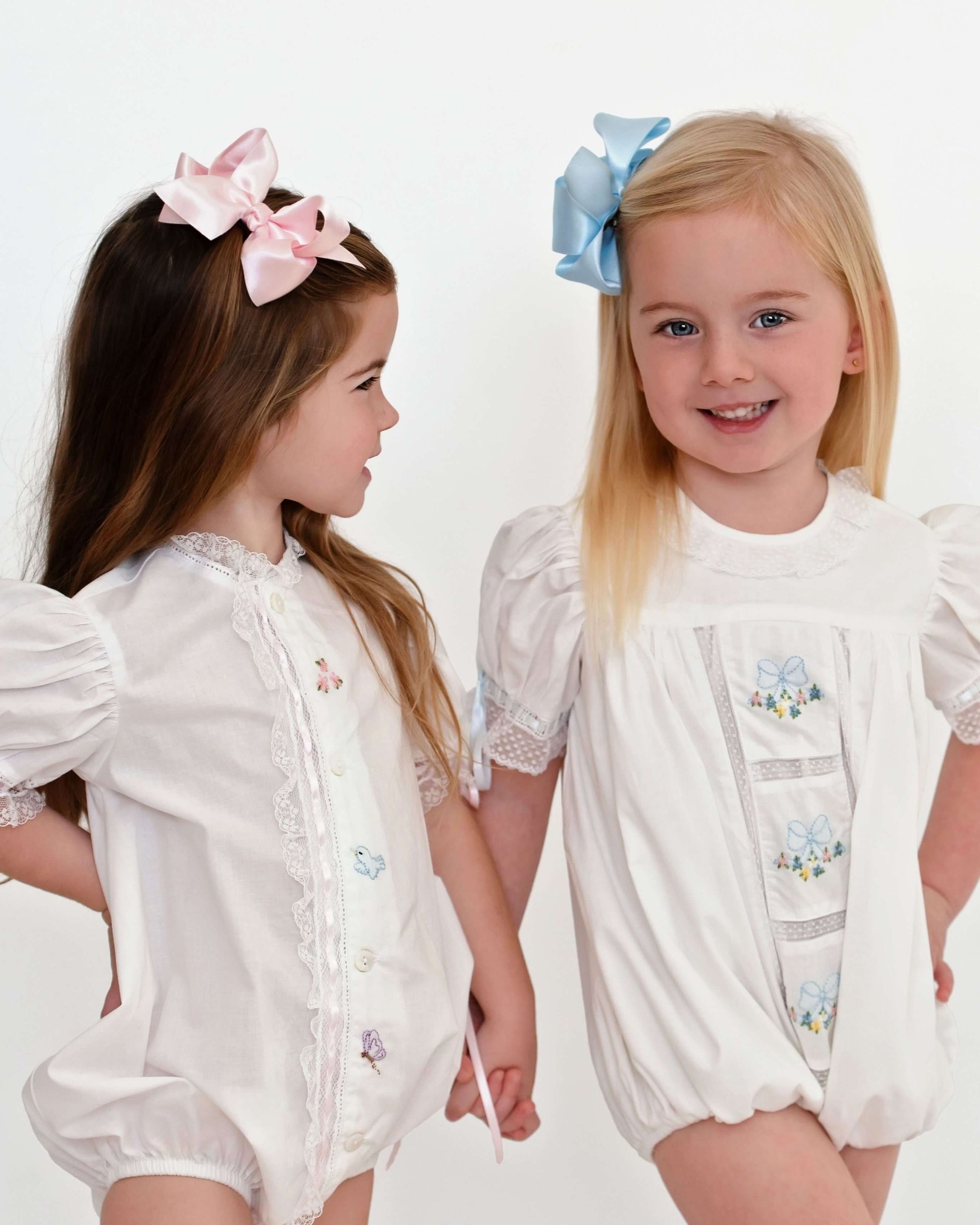 Two young girls wearing white outfits with decorative elements on a plain background