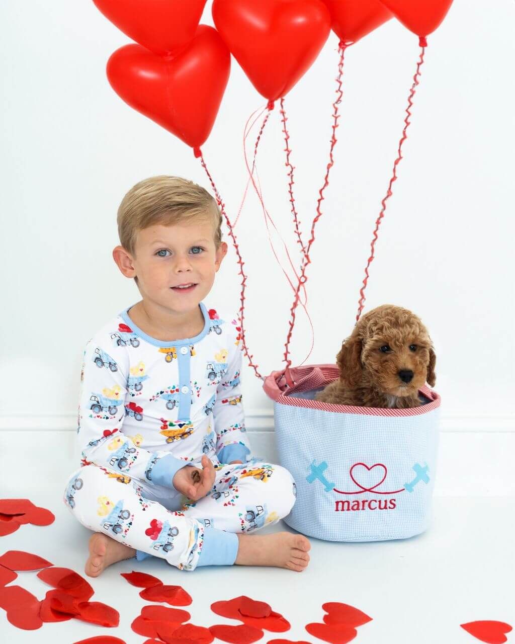 Child in pajamas with a small dog in a basket labeled 'marcus' surrounded by red heart balloons and confetti on a light background.