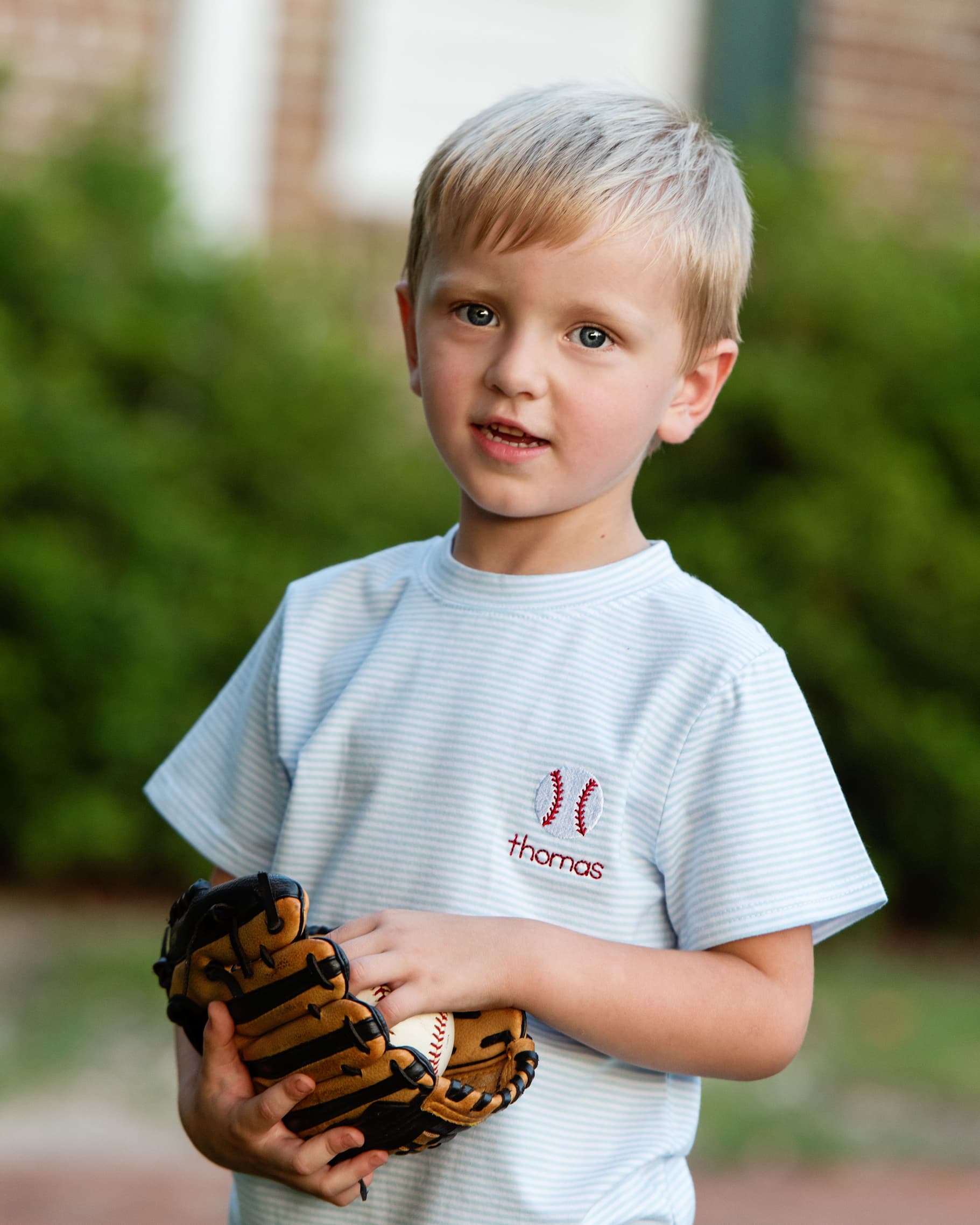 Young boy holding a baseball glove and ball, wearing a shirt with 'thomas' branding.