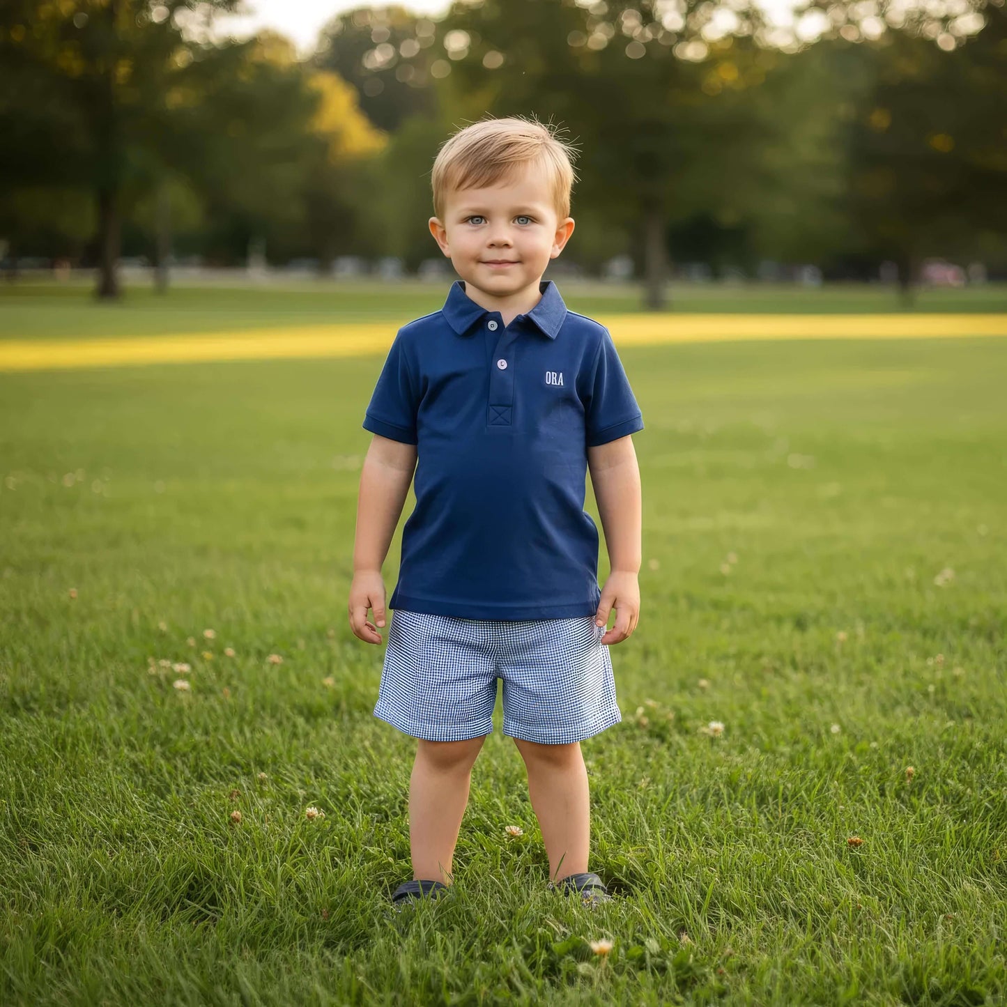Young boy in a blue shirt and shorts standing on a grassy field with trees in the background