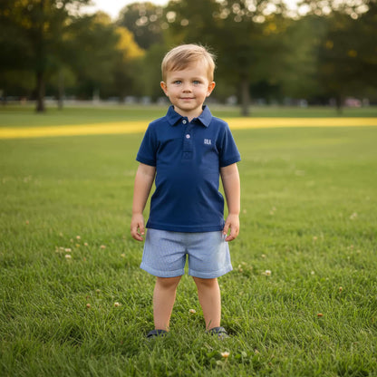 Young boy in a blue shirt and shorts standing on a grassy field with trees in the background