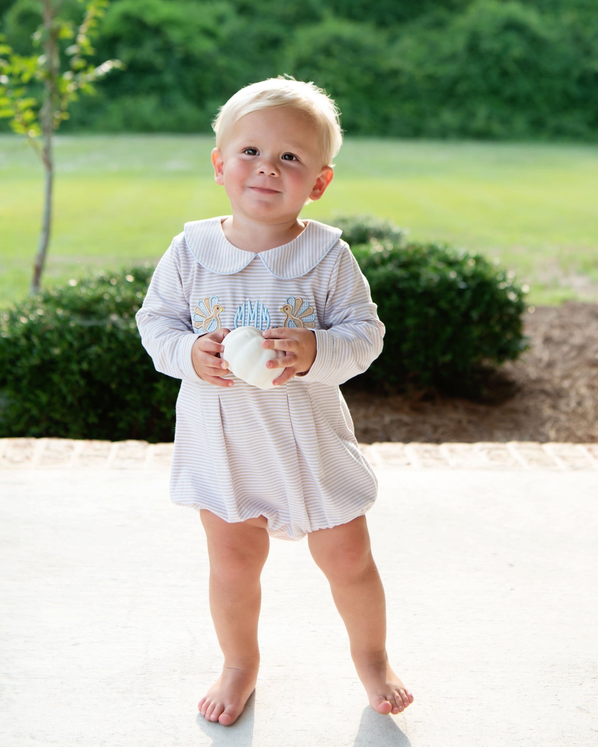 Young boy in a bubble with turkeys holding a white pumpkin outdoors with greenery in the background