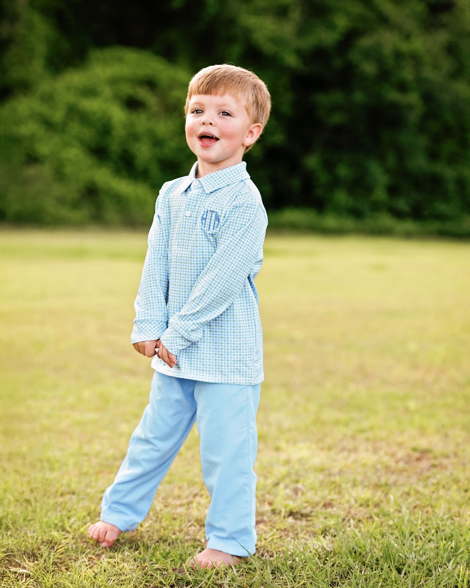 Young boy in a light blue checkered shirt and pants standing in a grassy field.