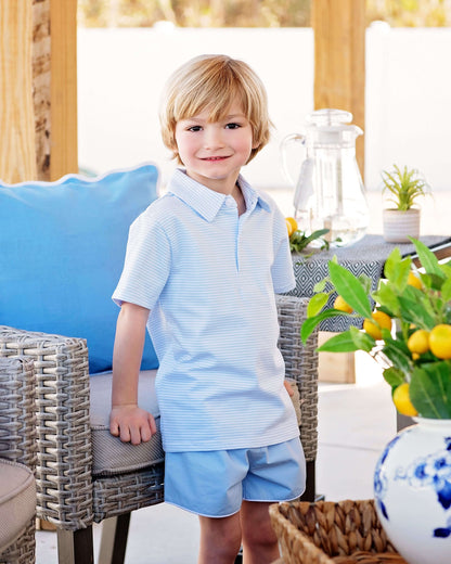 Young boy in a light blue outfit standing in an outdoor setting with furniture and plants.