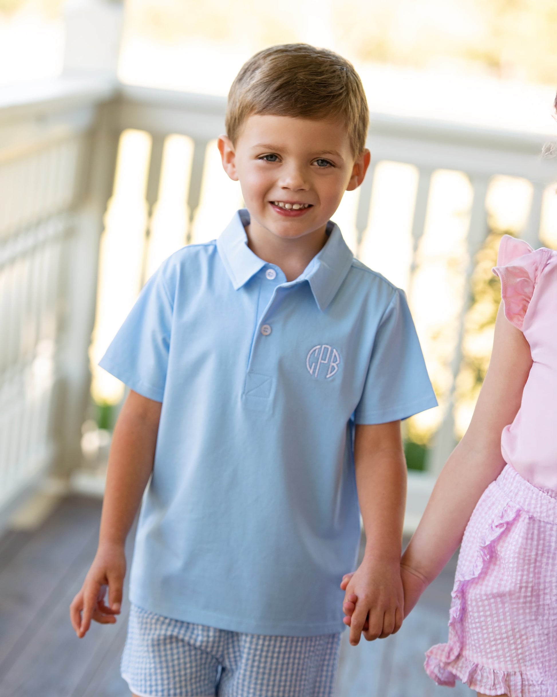 Young boy in a light blue polo polo with a monogram, holding hands with a girl in a pink dress on a wooden deck.