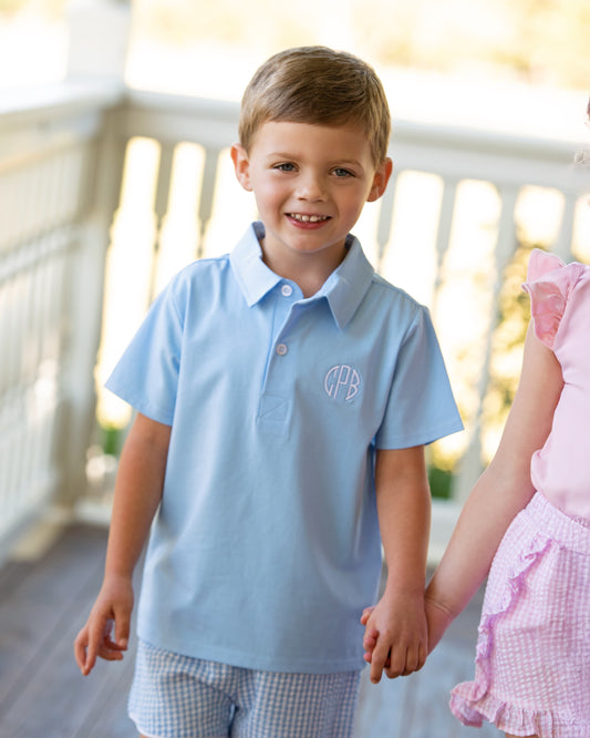 Young boy in a light blue polo polo with a monogram, holding hands with a girl in a pink dress on a wooden deck.