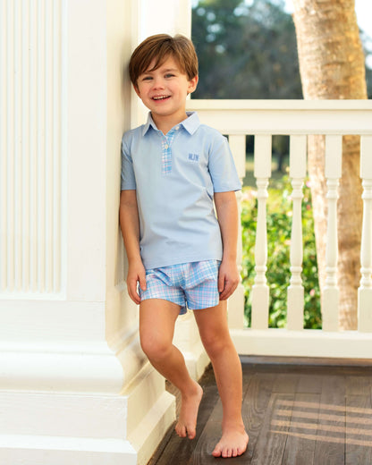 Young boy in a light blue polo shirt and plaid shorts standing on a staircase.