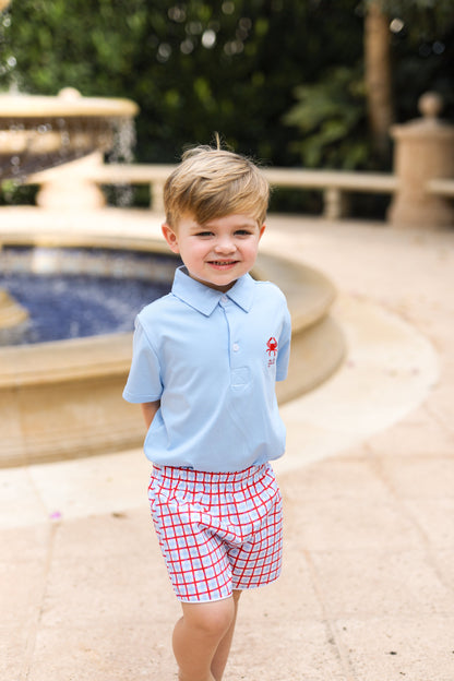 Young boy in a light blue polo shirt and red checkered shorts standing by a pool.