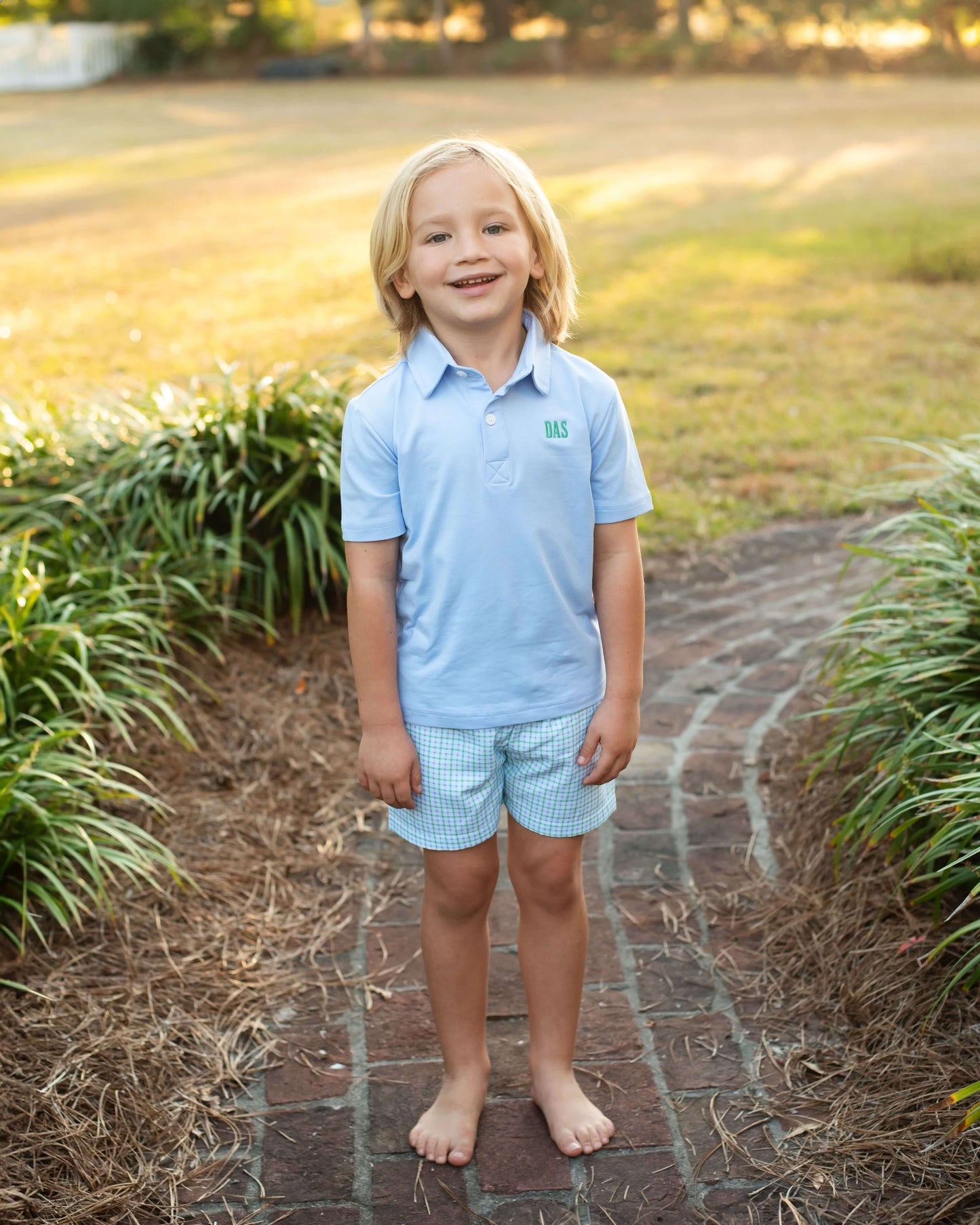 Young boy in a light blue polo shirt and shorts standing on a brick path with greenery around.