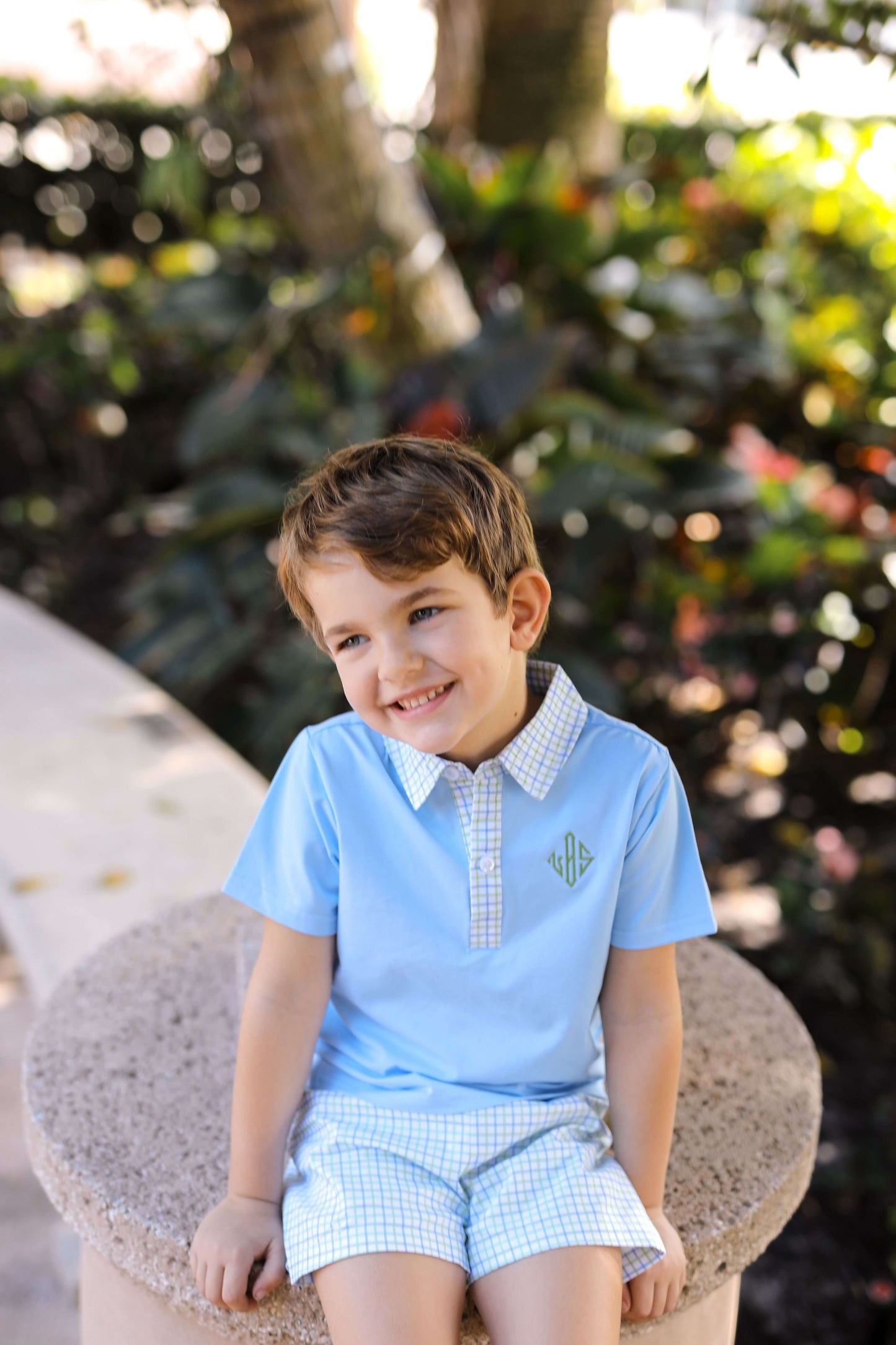 Young boy in a light blue shirt and checkered shorts sitting on a stone bench with greenery in the background.