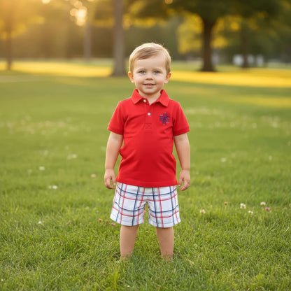Young boy in a red shirt and plaid shorts standing on a grassy field.