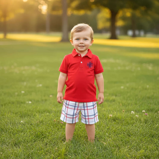 Young boy in a red shirt and plaid shorts standing on a grassy field.