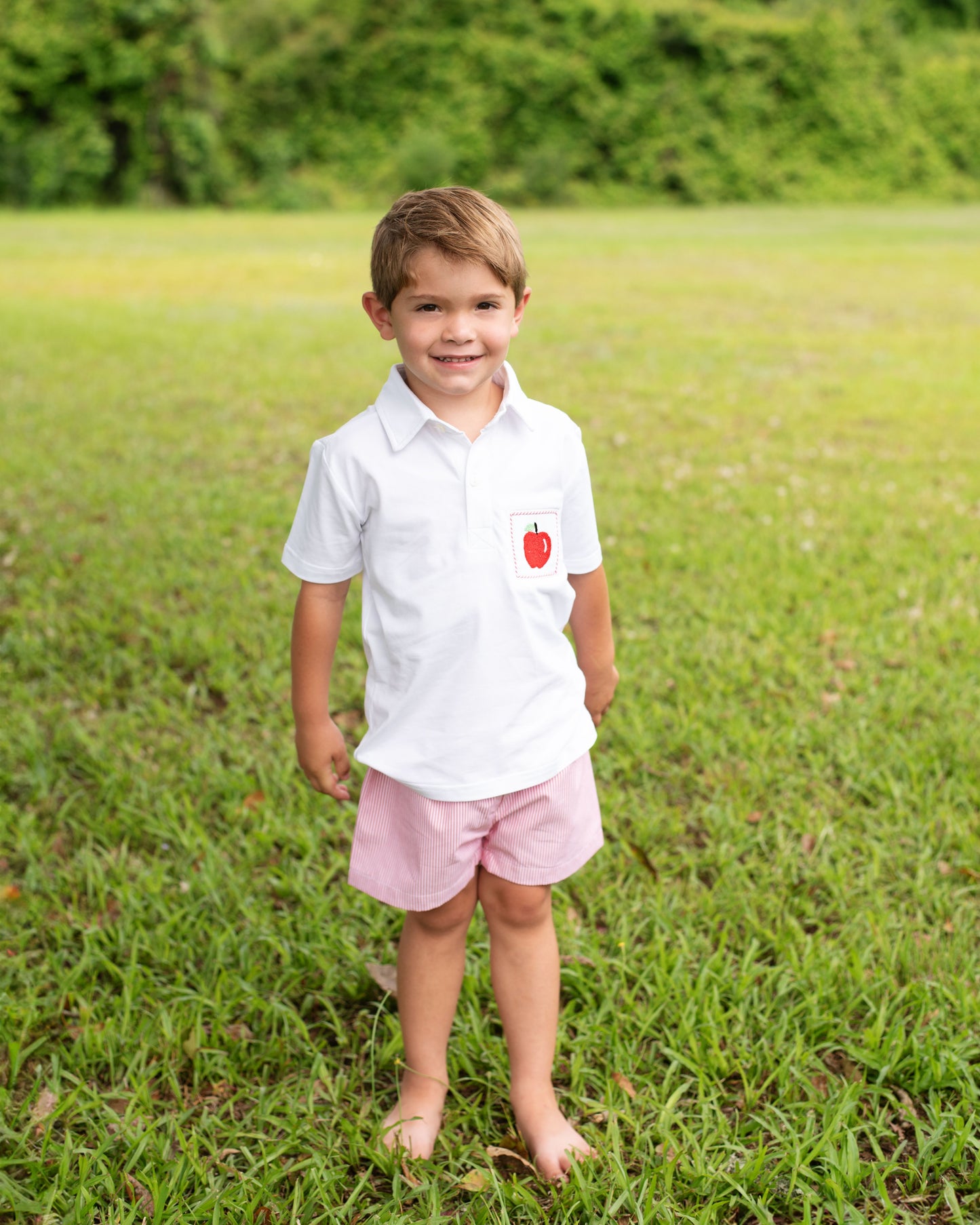 Young boy in a white shirt with an apple emblem and pink shorts standing in a grassy field.