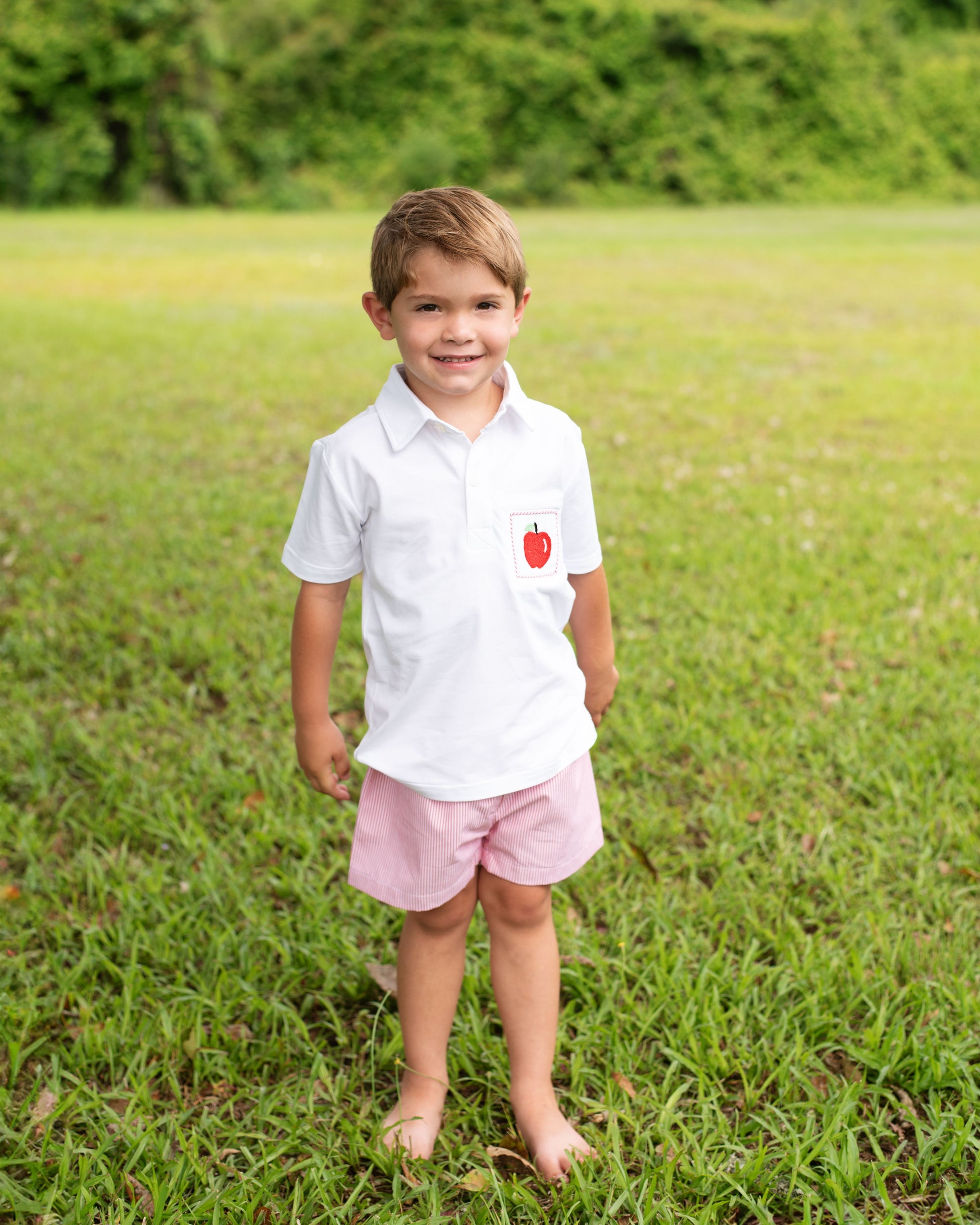 Young boy in a white shirt with an apple emblem and pink shorts standing in a grassy field.