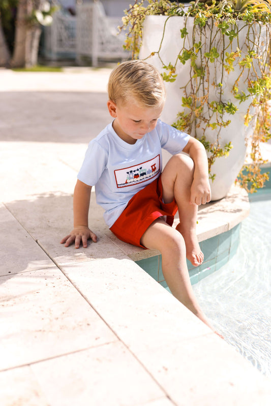 Young boy sitting by a pool wearing a light blue shirt with a graphic and red shorts.