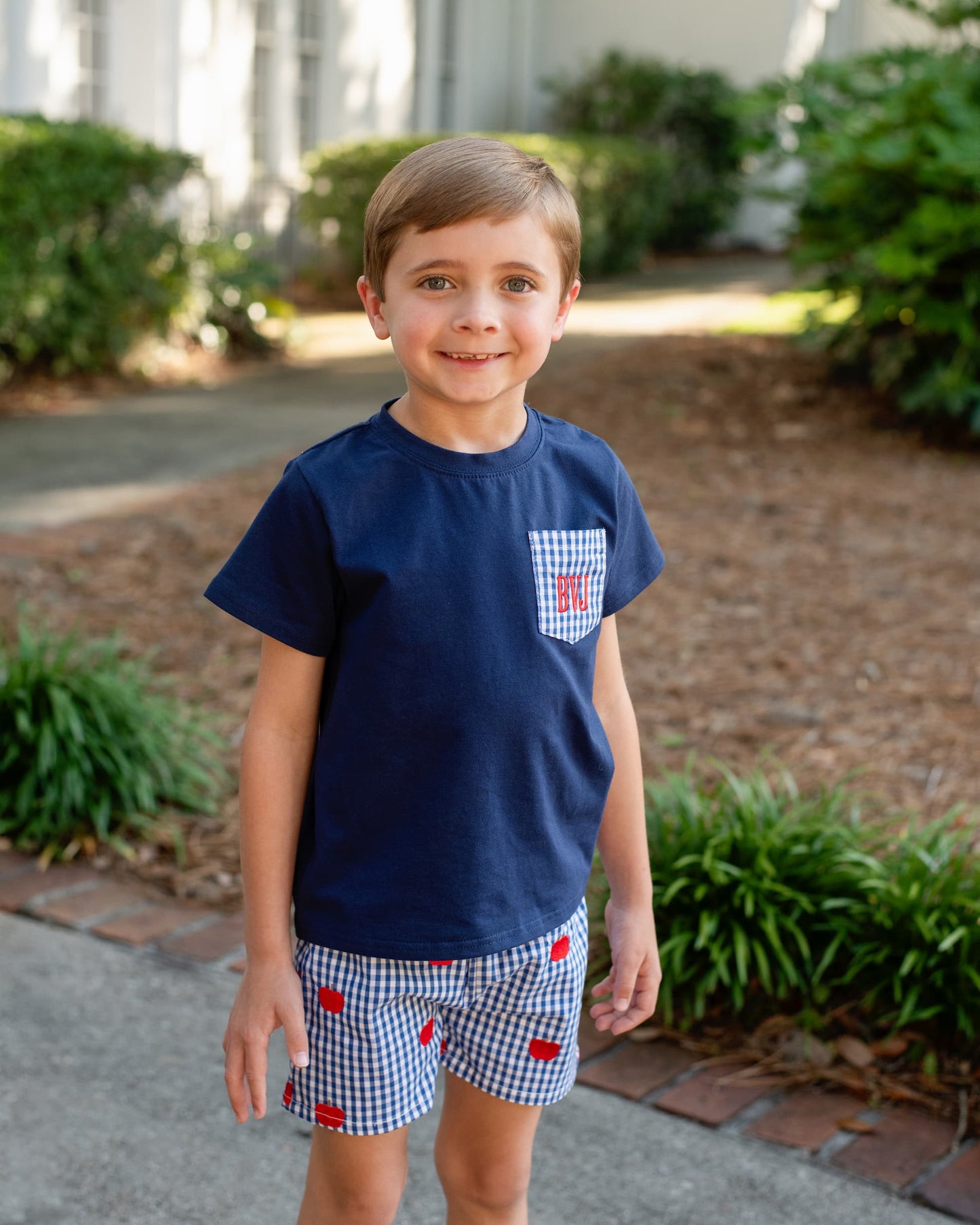 Young boy smiling wearing a navy shirt and navy gingham shorts with apples embroidered.
