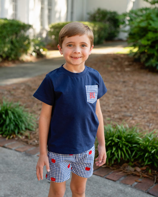 Young boy smiling wearing a navy shirt and navy gingham shorts with apples embroidered.