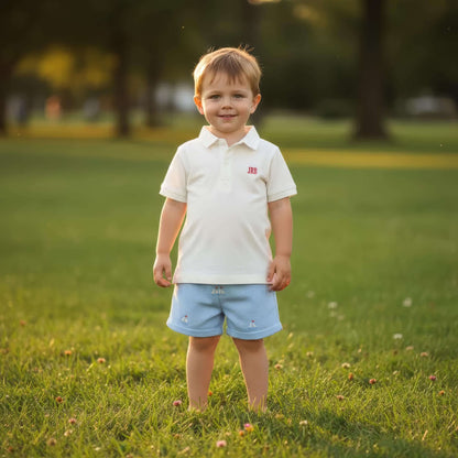 Young boy standing in a grassy field wearing a white polo shirt and light blue shorts.