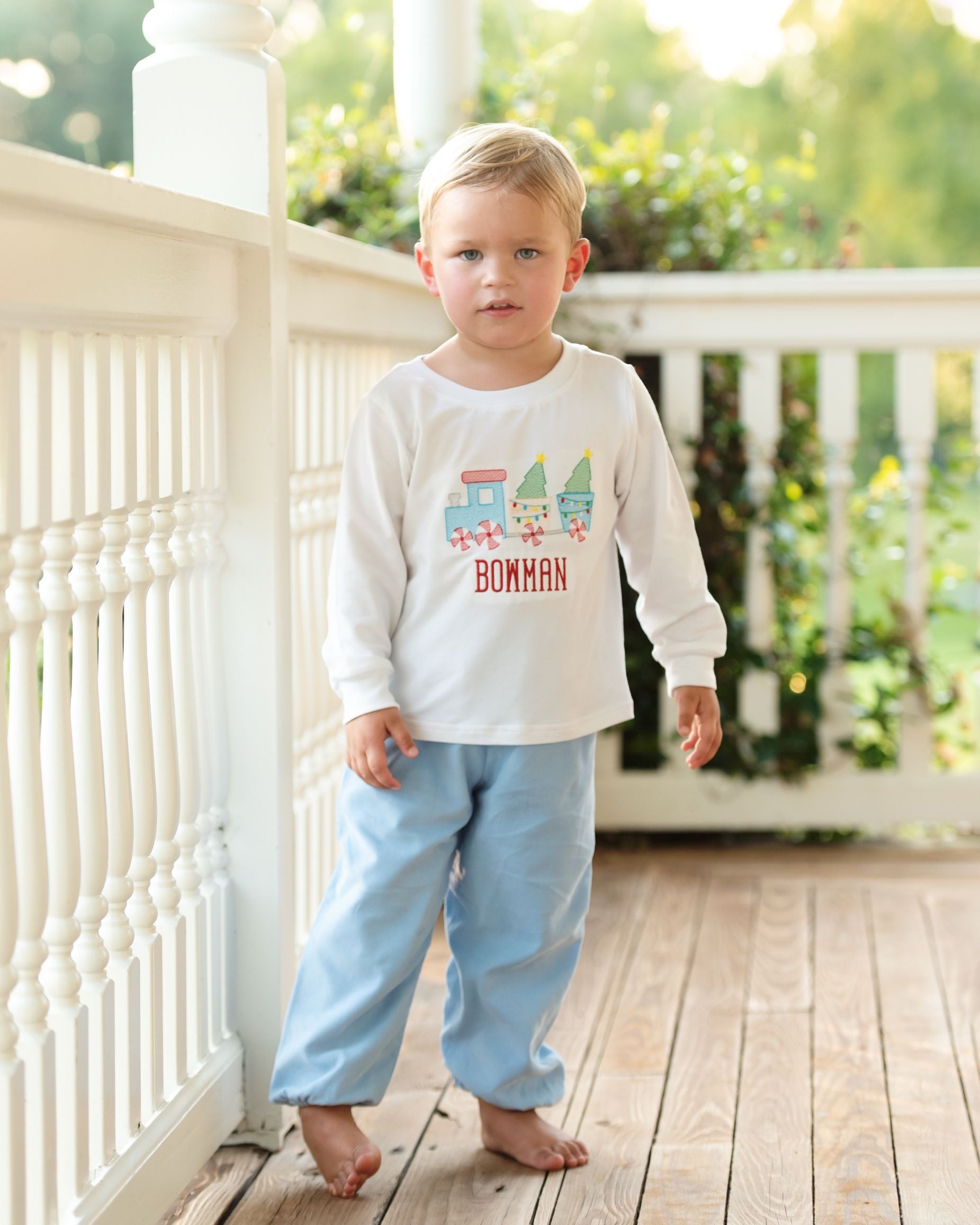 Young boy standing on a wooden deck wearing a white shirt with colorful train design and blue pants.