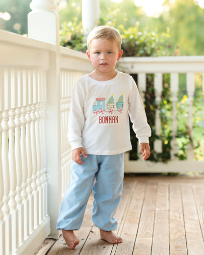Young boy standing on a wooden deck wearing a white shirt with colorful train design and blue pants.