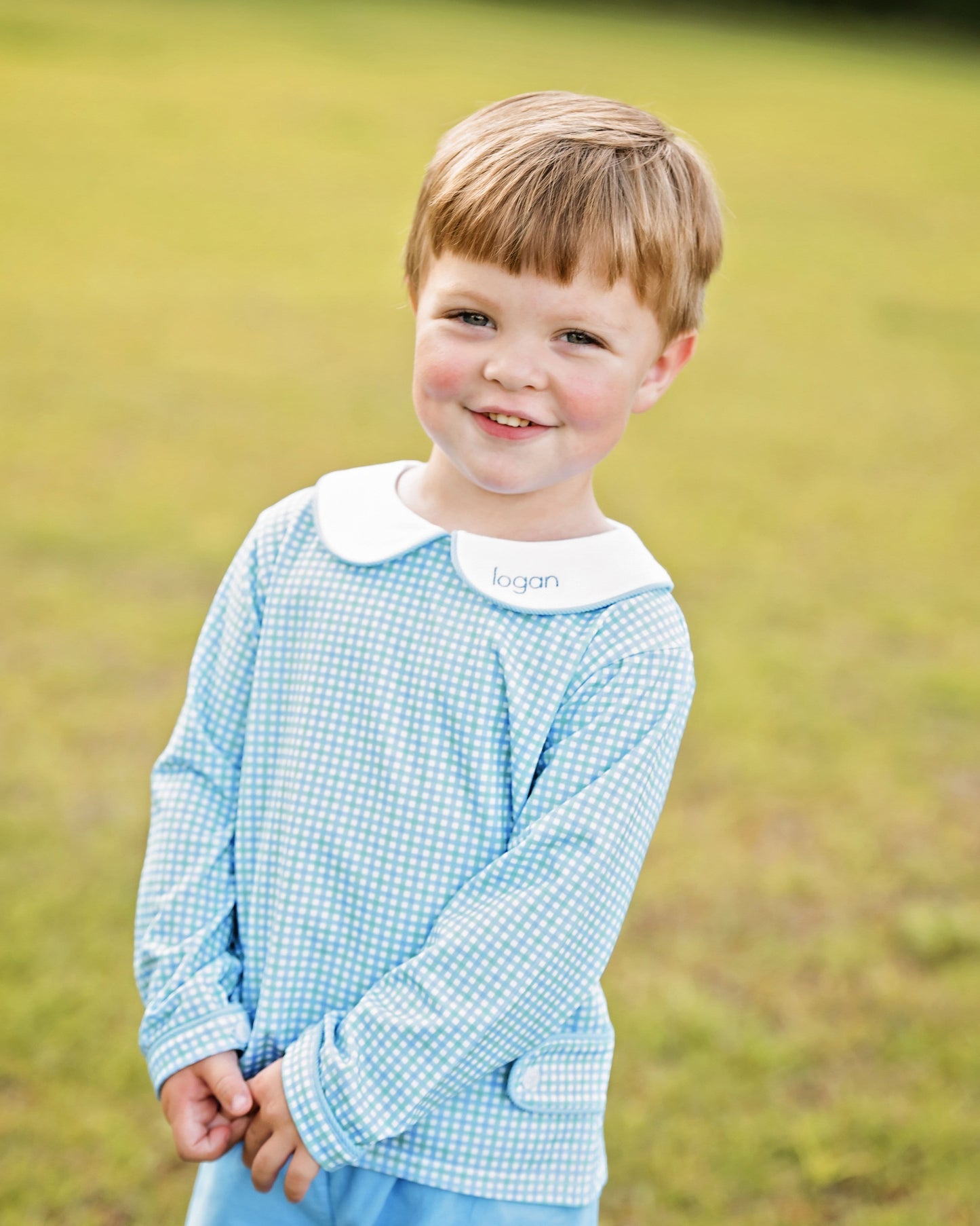 Young boy wearing a blue checkered shirt with a white collar in a grassy field