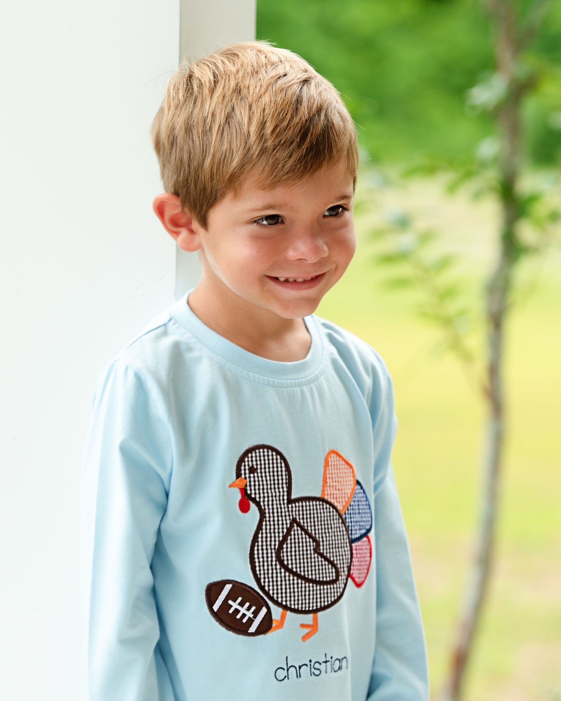 Young boy wearing a blue long sleeve with a colorful turkey design outdoors.