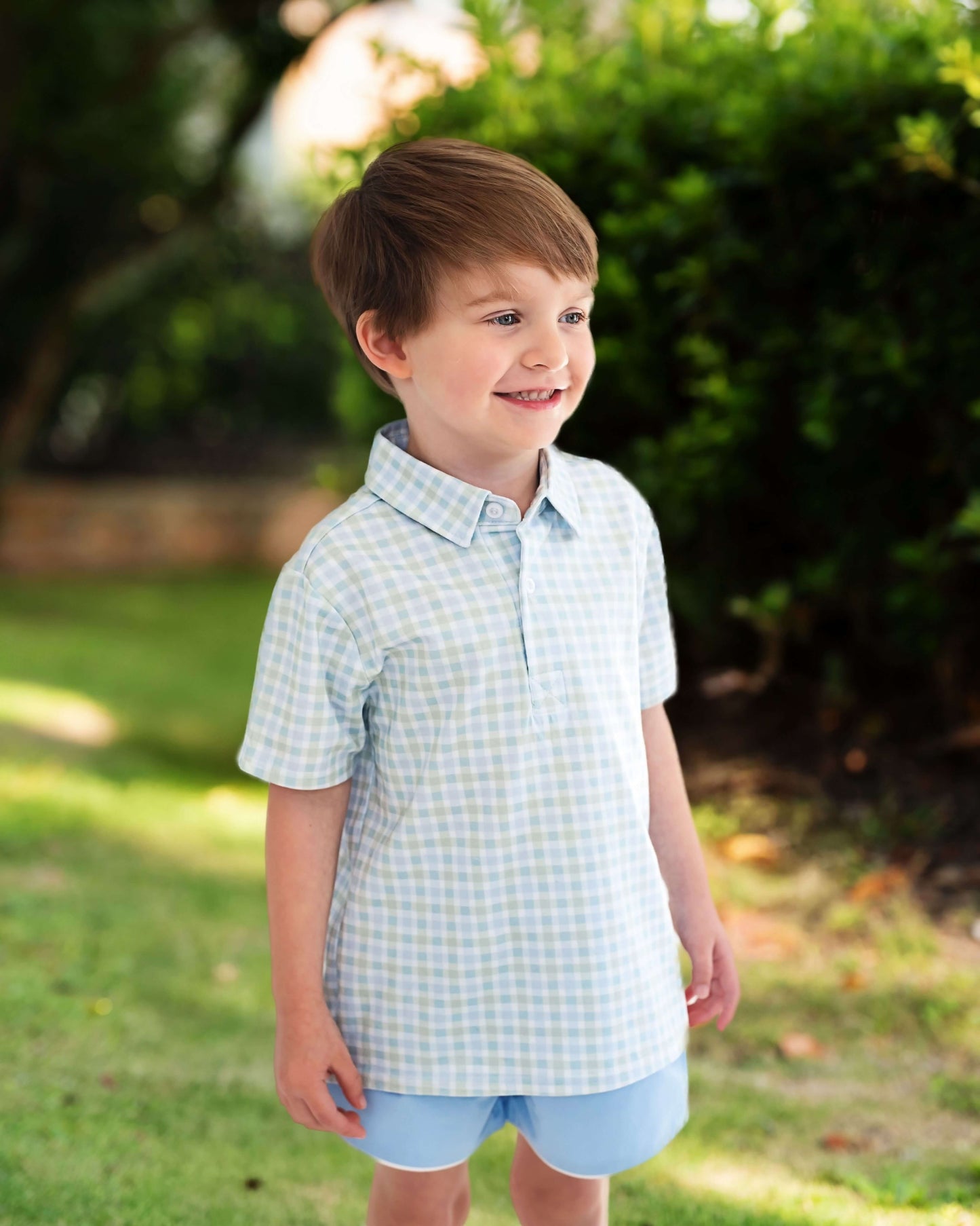 Young boy wearing a checkered shirt and shorts standing outdoors with greenery in the background