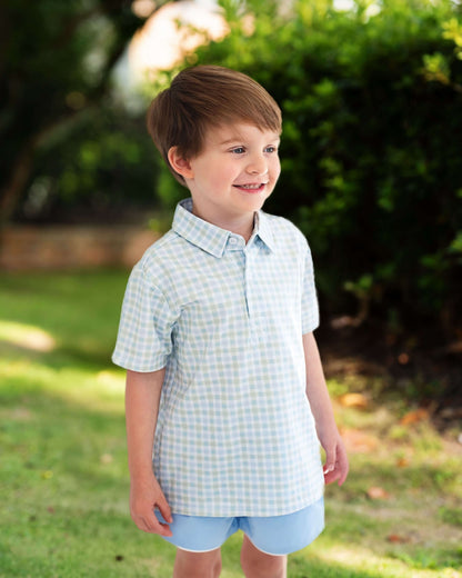 Young boy wearing a checkered shirt and shorts standing outdoors with greenery in the background