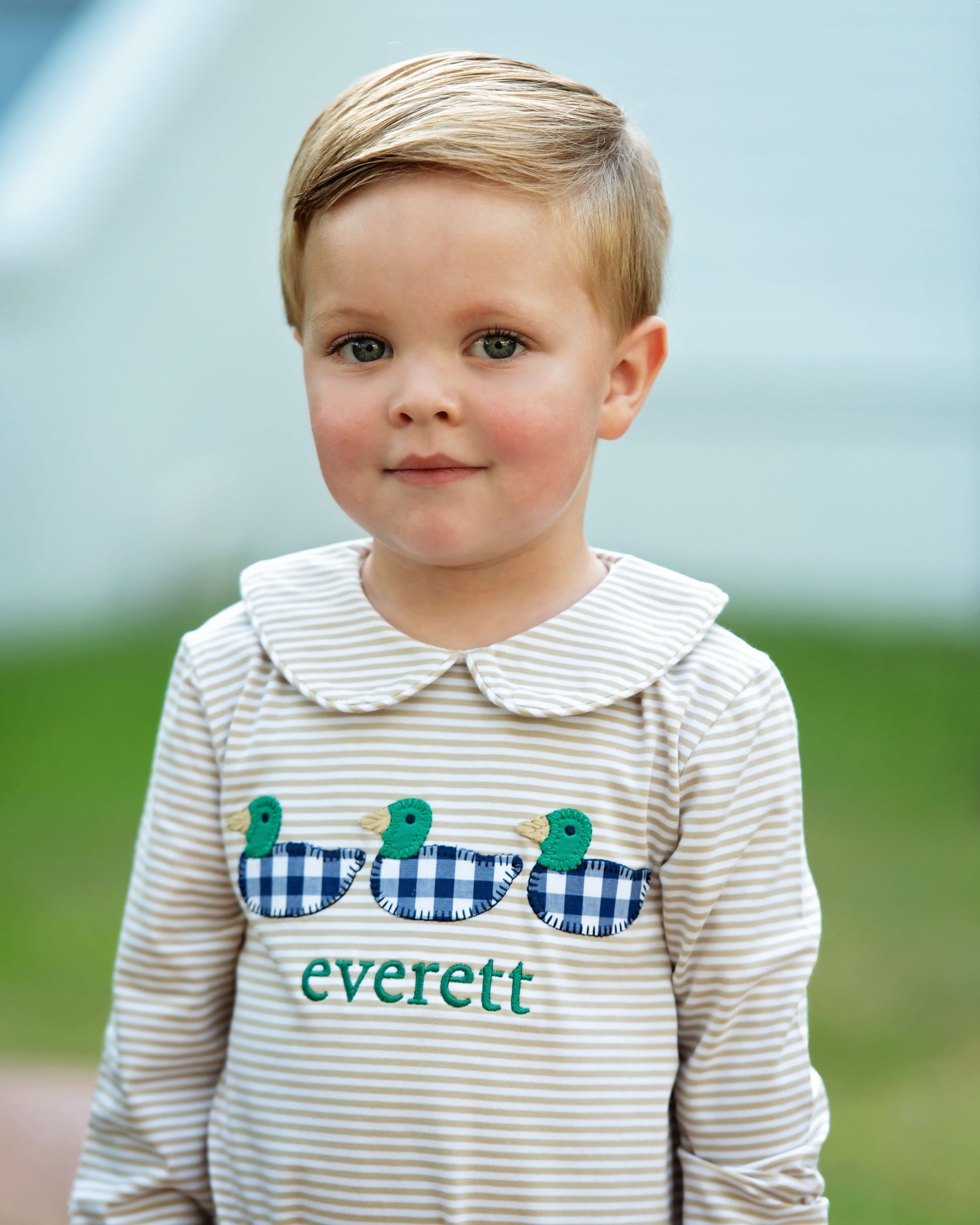 Young boy wearing a khaki striped shirt with a collar and duck design, standing outdoors.