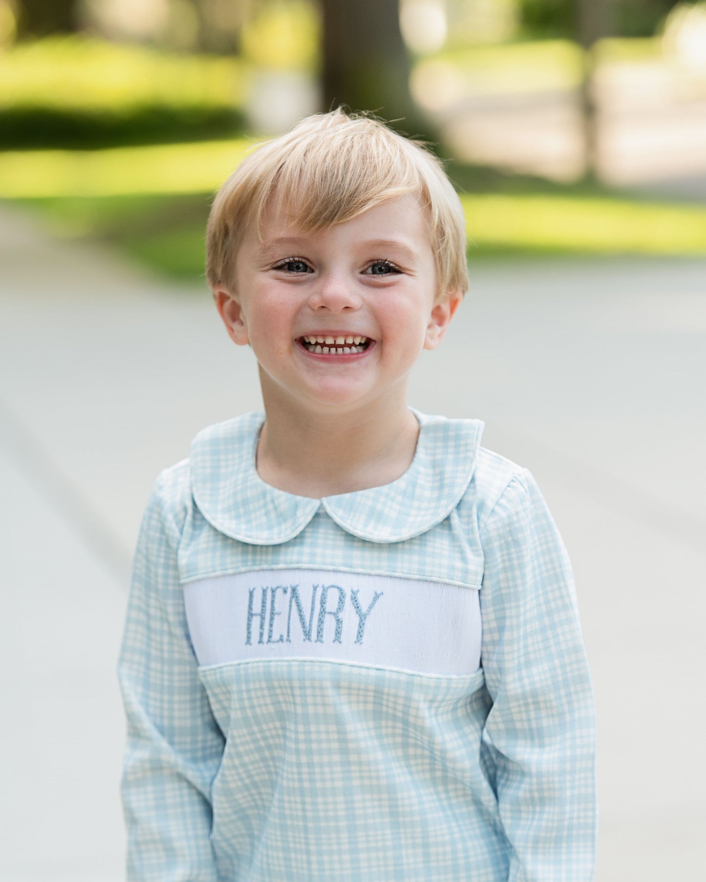 Young boy wearing a light blue checkered shirt with 'HENRY' embroidered on it, standing outdoors.