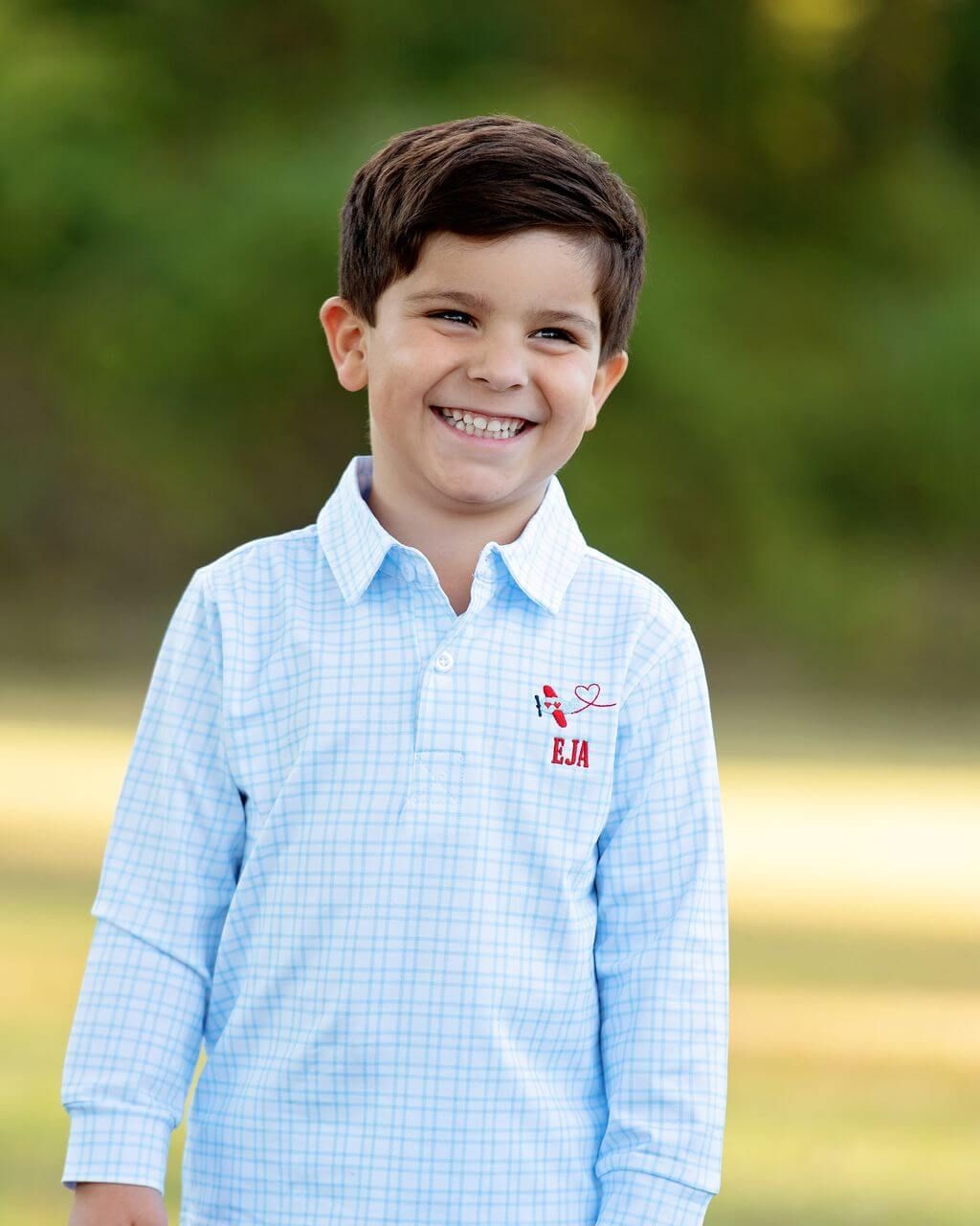 Young boy wearing a light blue checkered shirt with a logo, standing outdoors.