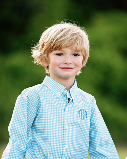 Young boy wearing a light blue checkered shirt with a monogram against a blurred green background