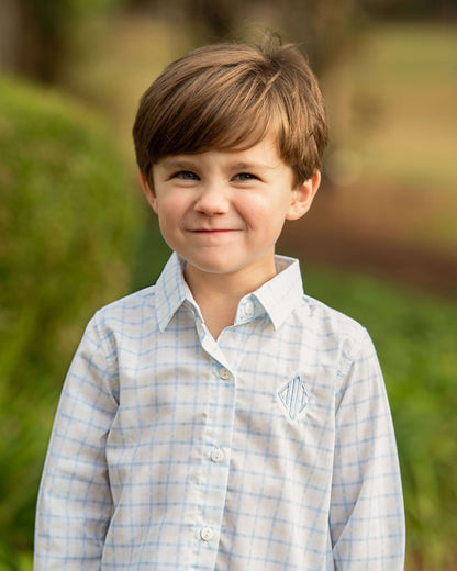 Young boy wearing a light blue checkered shirt with a monogram, standing outdoors with greenery in the background.